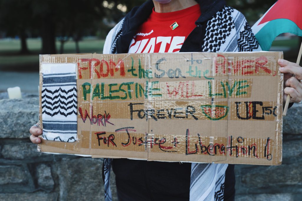 A person holds a sign that reads, "From the sea to the river, Palestine will live forever. The UE150 union logo is hand drawn on the side next to a watermelon. A kuffiyeh pattern adorns the left side of the side, while the bottom reads, "Work for justice and liberation!" The protestor waves a Palestinian flag in the other hand. They are standing in front of an old stone wall, topped with candles to honor the martyrs of the ongoing genocide.