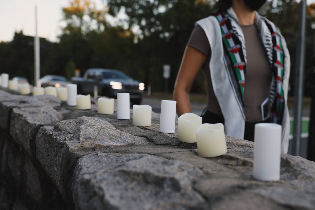 Candles adorn the top of an old stone wall. On the other side of the wall, a person walks along the sidewalk, a kuffiyeh with the Palestinian flag draped across her shoulders. In the street behind her, vehicles pass by, some honking their horns in support of Palestine.